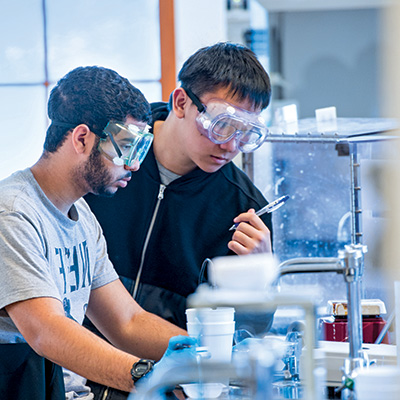 Two male student wearing safety goggles work together in a lab